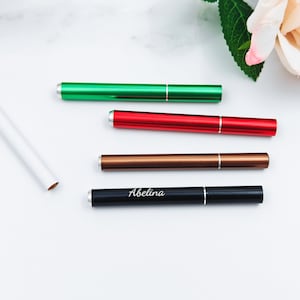 May include: Four brightly coloured cigarette cases in green, red, brown, and black, with a cigarette on the left. The black case has the name "Abelina". The cases are on a white surface.