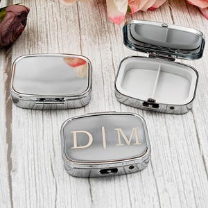 Puede incluir: Tres pastilleros rectangulares plateados, uno abierto, que muestran un interior blanco con dos compartimentos. Una caja tiene las iniciales "D|M" en la tapa. Las cajas están sobre una superficie de vetas de madera clara, con flores en el fondo.
