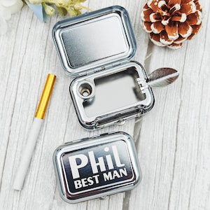Op de afbeelding: Een zilveren metalen doos met de tekst "Phil BEST MAN" in het wit. De doos is open en onthult een klein compartiment en een metalen blad-vormig stuk. Een goud-witte sigaret ligt naast de doos. De doos staat op een licht houten oppervlak.
