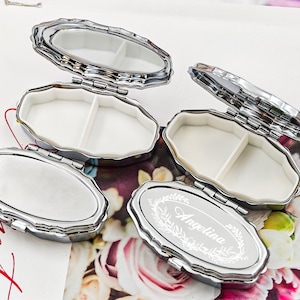 May include: Silver-toned, oval pill boxes with hinged lids, some open to reveal white interiors divided into two compartments. One box is engraved with the name "Angelina" in a decorative font. The boxes are arranged on a floral background.