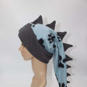 May include: A blue and gray fleece hat with a long tail and black paw print design. The hat has a pointed, spiky tail.
