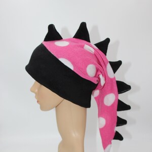 May include: A pink and black fleece hat with white polka dots and a long, black tail. The hat has a pointed top and is shaped like a dragon's head.