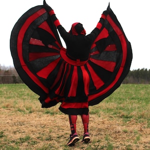 Könnte beinhalten: Eine Person trägt einen schwarzen und roten Kapuzenumhang mit einem großen kreisförmigen Design. Der Umhang weht im Wind und die Person steht auf einem Feld.