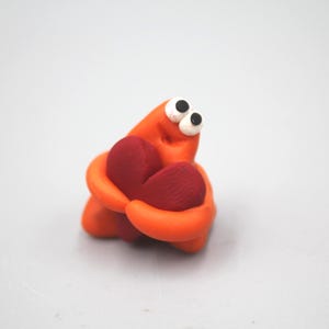 May include: A whimsical orange clay figure with large white eyes and black pupils, embracing a red heart. The figure is set against a plain white background, creating a simple, yet endearing composition.