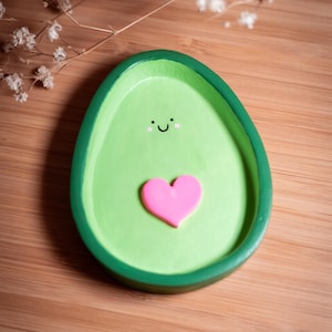 Pode incluir: Uma bandeja verde em forma de abacate com um coração rosa e um rosto sorridente. A bandeja está sobre uma superfície de madeira. A bandeja de abacate é um item decorativo.