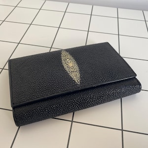 May include: A black leather wallet with a silver-colored stingray skin design. The wallet is folded in half and has a smooth, textured surface.