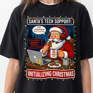 Peut inclure: Un t-shirt noir avec une illustration du Père Noël portant un casque et travaillant sur un ordinateur portable. Le texte indique SANTA'S TECH SUPPORT: 404 CHRISTMAS NOT FOUND, OFFICIAL NORTH SNOWDOME IT SPECIALIST et INITIALIZING CHRISTMAS, entouré de guirlandes lumineuses.