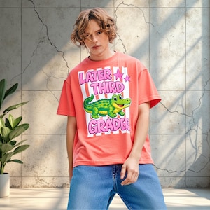 Könnte beinhalten: Korallfarbenes T-Shirt mit dem Text "Later Third Grade" und einer Cartoon-Alligator-Grafik. Das Shirt hat einen Rundhalsausschnitt und kurze Ärmel. Die Person trägt eine blaue Jeans.