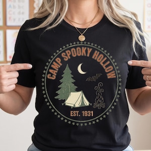 May include: A black short-sleeved t-shirt featuring a circular graphic with the text "CAMP SPOOKY HOLLOW" and "EST. 1931". The design includes a green pine tree, a white tent, a crescent moon, a flying bat, and a campfire, all enclosed by stars.
