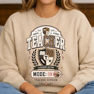 Peut inclure: Un sweat-shirt à col rond beige avec un motif graphique incluant le texte TEACHER, CLASSROOM EDITION, MODE: ON et TEACHER DIVISION. Le design présente un écusson à motif léopard, un livre, un crayon et une icône en forme d'étoile.