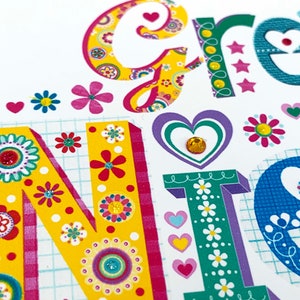 May include: Close-up of a colorful greeting card with the word "GREETINGS" in large, decorative letters. The letters are adorned with floral patterns, hearts, and sparkling embellishments. The background is white with a grid pattern.