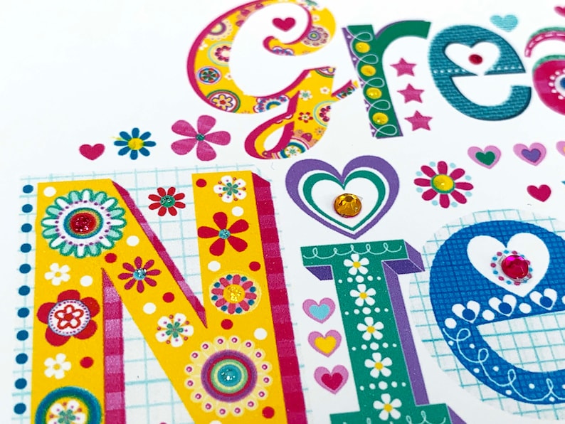 May include: Close-up of a colorful greeting card with the word "GREETINGS" in large, decorative letters. The letters are adorned with floral patterns, hearts, and sparkling embellishments. The background is white with a grid pattern.