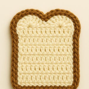 May include: A crocheted slice of bread with a light beige center and a brown crust. The handmade bread slice has a textured appearance, with visible stitch patterns. The crust is a darker brown, creating a defined border around the bread.