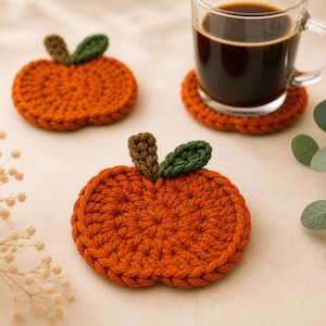 May include: Three orange crochet pumpkin coasters with brown stems and green leaves. One coaster holds a glass mug filled with dark coffee. The coasters are handmade and sit on a neutral surface, perfect for fall decor.