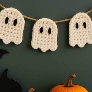 May include: A Halloween-themed image featuring a string of cream-colored, crocheted ghost decorations with black eyes. The ghosts are strung on a natural twine. The background includes orange pumpkins, black bat and witch hat cutouts, and a dark green wall.