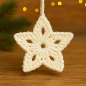 Puede incluir: Adorno de estrella de ganchillo de color crema, con un diseño de cinco puntas y detalles calados. La estrella está suspendida por un hilo blanco, ideal para la decoración navideña. El fondo presenta una superficie de madera borrosa y luces bokeh.