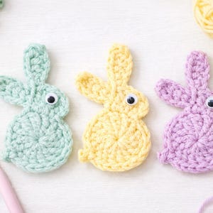 May include: Three pastel-coloured crochet bunny decorations: mint green, yellow, and lavender. Each bunny has googly eyes. Yarn and a crochet hook are also visible, suggesting a crafting theme.