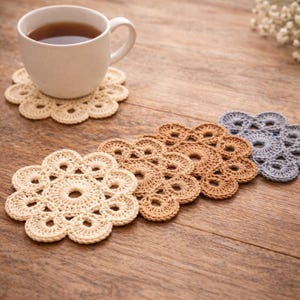 May include: A white ceramic mug with tea sits on a beige crocheted coaster. Several flower-shaped coasters in beige, brown, and gray are arranged on a wooden surface. The coasters are handmade and have a delicate, openwork design.