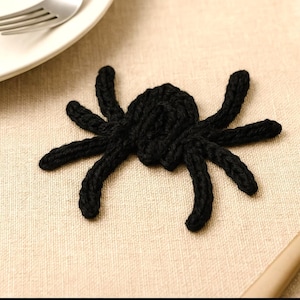 May include: A black, knitted spider decoration with eight legs. The spider is made of a thick, textured yarn and is placed on a beige surface. The spider is a Halloween or holiday-themed craft item.
