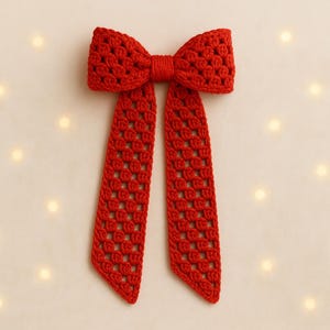 May include: A handmade red crochet bow with long, flowing tails. The bow features a textured, open weave pattern and is displayed against a soft, off-white background with scattered, glowing lights. This decorative bow is suitable for various craft projects.