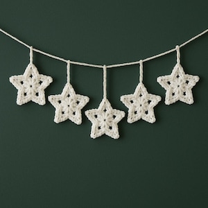 May include: A garland of five white crocheted stars suspended from a white string. Each star is a five-pointed shape with a detailed openwork design. The background is a solid, dark green color.