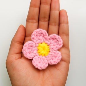 May include: A handmade crochet flower with pink petals and a yellow center. The flower is held in an open hand against a white background. The crochet stitches are visible, giving the flower a textured appearance.