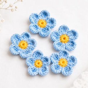 May include: Five handmade crochet flowers in a light blue with yellow centres. The flowers are arranged in a circular pattern on a white surface. The petals are detailed with visible stitching.