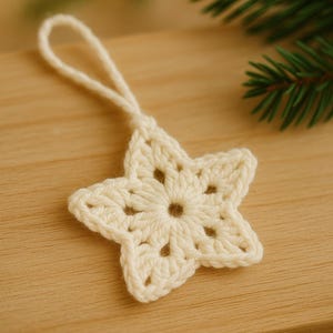 May include: Cream-colored, crocheted star ornament with a loop for hanging. The star has a five-point design with openwork details. The ornament is displayed on a wooden surface, with a sprig of evergreen in the background. A festive, handmade item.