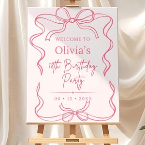 May include: A white canvas sign on a wooden easel, featuring pink text and decorative ribbon designs. The sign reads "Welcome to Olivia's 18th Birthday Party" with a date. The easel is made of light-colored wood.