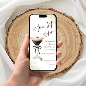 May include: A smartphone displays a birthday invitation with a cocktail illustration. The text reads "a tini bit older" above the invitation details, including the date, time, and location of the party. The invitation features a martini glass with a black bow.