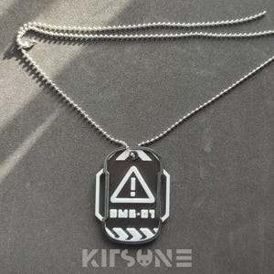 May include: A black and white dog tag pendant with a silver chain. The tag features a white exclamation point inside a triangle, the text "UMB-01", and a series of white arrows. The word "KITSUNE" is at the bottom of the tag.