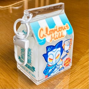 Op de afbeelding: Transparante sleutelhanger in de vorm van een melkpak met een blauw-wit gestreept dakontwerp. Het pakje heeft de tekst "Glorious Milk" en een stripfiguur met een glas melk. Inclusief een zilverkleurige sluiting en een QR-code.