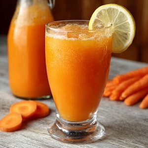 Könnte beinhalten: Ein Glas frischer Karottensaft mit einer Zitronenscheibe als Garnierung. Eine volle Glasflasche Karottensaft befindet sich hinter dem Glas. Auf dem Tisch liegen geschnittene Karotten. Der Saft hat eine leuchtend orange Farbe.