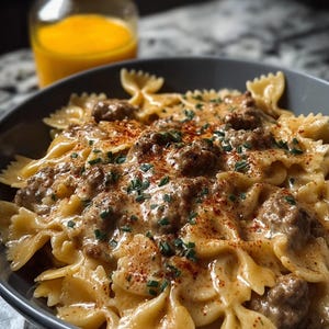 Pode incluir: Uma foto em close-up de uma tigela cinza cheia de massa farfalle cozida, coberta com um molho cremoso, carne moída e polvilhada com ervas e especiarias vermelhas. Um frasco de vidro com líquido laranja está no fundo.