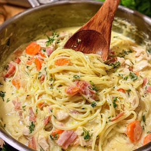 Könnte beinhalten: Eine Edelstahlpfanne gefüllt mit cremiger Pasta. Das Gericht enthält Spaghetti, Hühnchen, Karotten und Speck, wobei ein Holzlöffel die Zutaten verrührt. Frische Kräuter garnieren das Gericht.