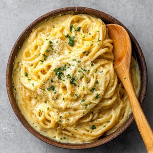 Könnte beinhalten: Draufsicht auf eine Holzschüssel mit cremiger Pasta. Die Pasta ist mit einer reichhaltigen, gelben Sauce überzogen und mit geriebenem Käse und frischen grünen Kräutern garniert. Ein Holzlöffel liegt in der Schüssel.