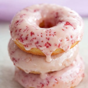 Könnte beinhalten: Ein Stapel von drei Erdbeer-Glasur-Donuts. Die Donuts haben eine hellrosa Glasur mit sichtbaren roten Erdbeerstücken. Die Glasur tropft an den Seiten der Donuts herunter, die übereinander gestapelt sind.