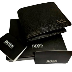 May include: Black leather wallet with the BOSS logo embossed on the front. The wallet is displayed with its original packaging, including a box and a card, all bearing the BOSS brand name. The wallet has a textured surface.