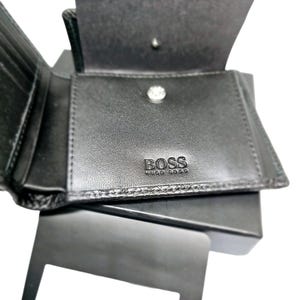 May include: Black leather wallet with the embossed brand name "BOSS" visible on the front. The wallet is open, revealing multiple card slots and a coin compartment. The wallet is sitting on a black box.