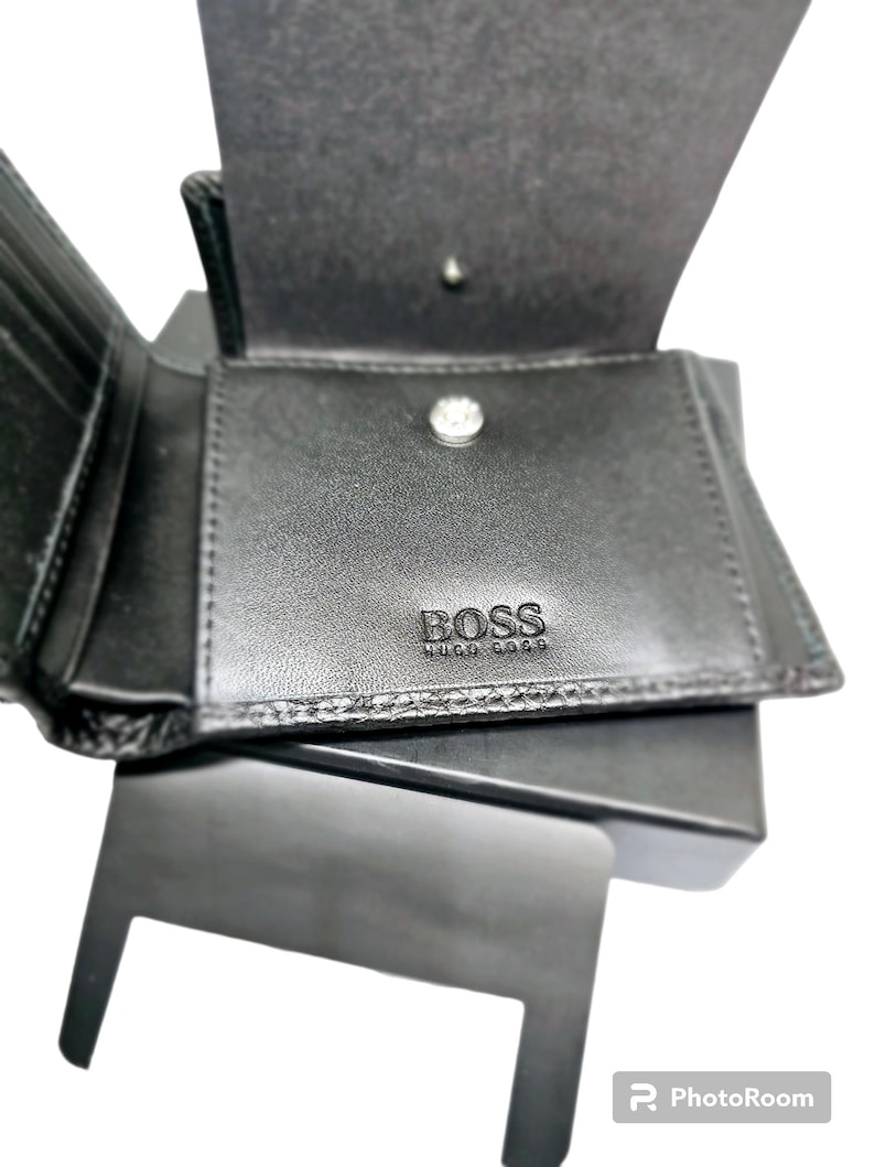 May include: Black leather wallet with the embossed brand name "BOSS" visible on the front. The wallet is open, revealing multiple card slots and a coin compartment. The wallet is sitting on a black box.