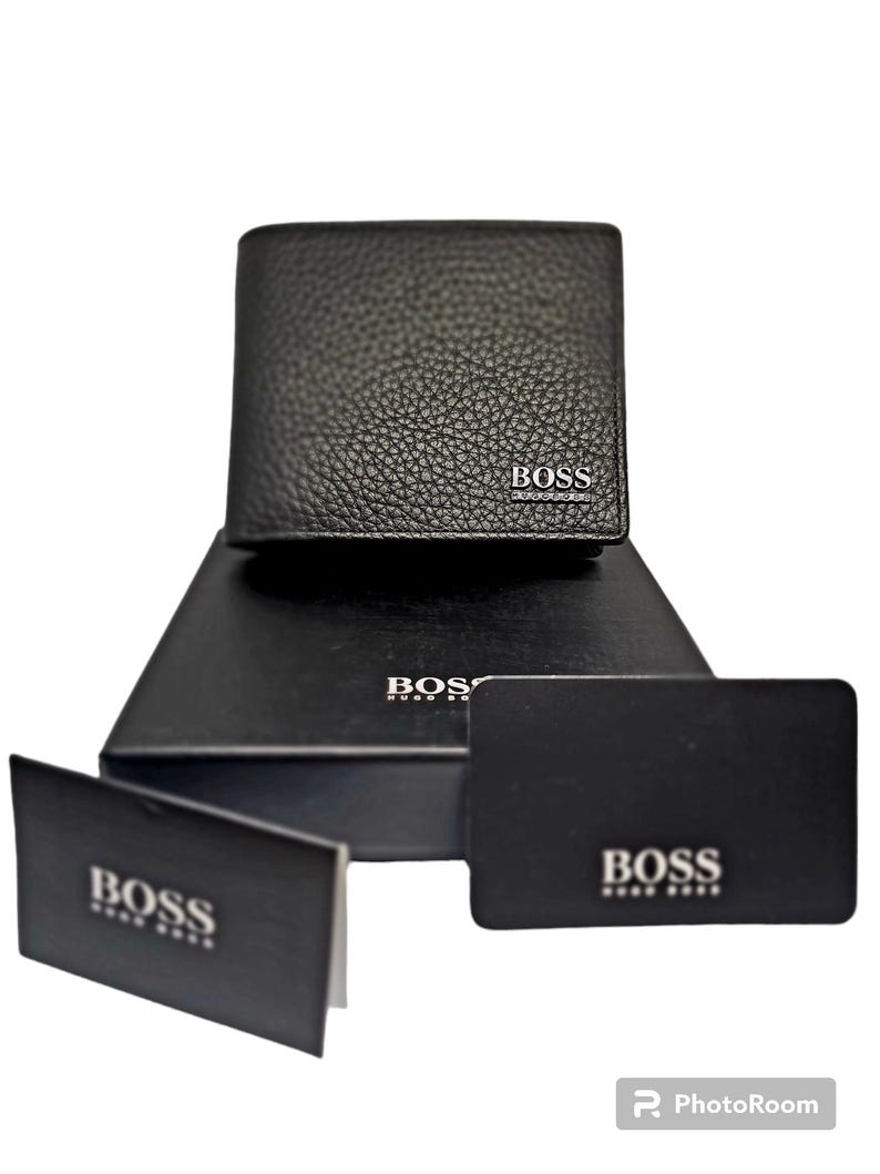 May include: Black leather wallet with the brand name "BOSS" in silver lettering. The wallet is displayed on a black box and accompanied by two black cards, also bearing the "BOSS" logo. The wallet has a textured surface.