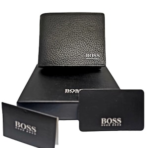 Op de afbeelding: Zwart leren portemonnee met de merknaam "BOSS" in zilveren letters. De portemonnee wordt gepresenteerd op een zwarte doos en vergezeld van twee zwarte kaarten, eveneens met het "BOSS"-logo. De portemonnee heeft een gestructureerd oppervlak.