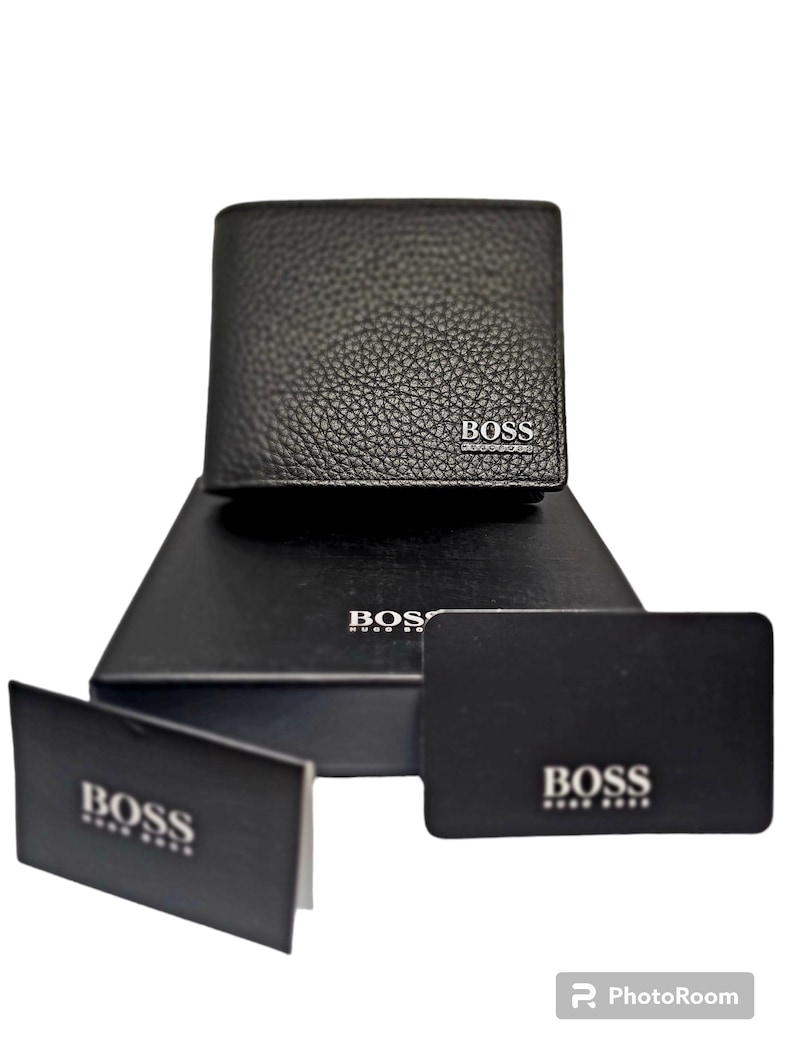 May include: Black leather wallet with the brand name "BOSS" in silver lettering. The wallet is displayed on a black box and accompanied by two black cards, also bearing the "BOSS" logo. The wallet has a textured surface.