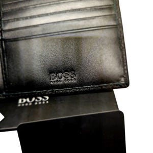 May include: Black leather wallet with the embossed brand name "BOSS". The wallet is open, revealing multiple card slots. A black card with the brand name "BOSS" is visible below the wallet.