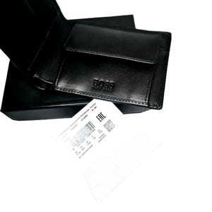 Pode incluir: Carteira de couro preta com o nome da marca "BOSS" em relevo na frente. A carteira est&aacute; aberta, revelando os compartimentos internos. Um pequeno bolso quadrado para moedas &eacute; vis&iacute;vel. Uma etiqueta branca com texto e um c&oacute;digo de barras est&aacute; em primeiro plano.
