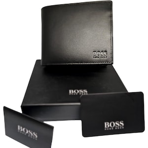 Puede incluir: Cartera de cuero negro con el nombre de la marca "BOSS" en relieve. Se muestra sobre una caja negra y acompañada de dos tarjetas negras, también con el logotipo "BOSS". La presentación general es elegante y minimalista.