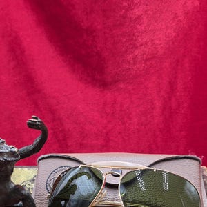 May include: Gold-framed aviator sunglasses with green lenses rest on a tan case. The sunglasses have a classic design with a double bridge and teardrop-shaped lenses. The case is rectangular and has a textured surface. The background is a deep red.