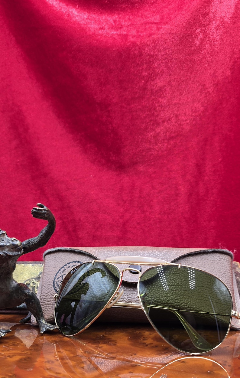 May include: Gold-framed aviator sunglasses with green lenses rest on a tan case. The sunglasses have a classic design with a double bridge and teardrop-shaped lenses. The case is rectangular and has a textured surface. The background is a deep red.