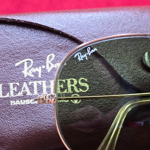 May include: Close-up of Ray-Ban sunglasses with a gold frame and dark lenses. The Ray-Ban logo is visible on the lens. A brown leather case with the Ray-Ban logo is in the background. The sunglasses are a classic aviator style.