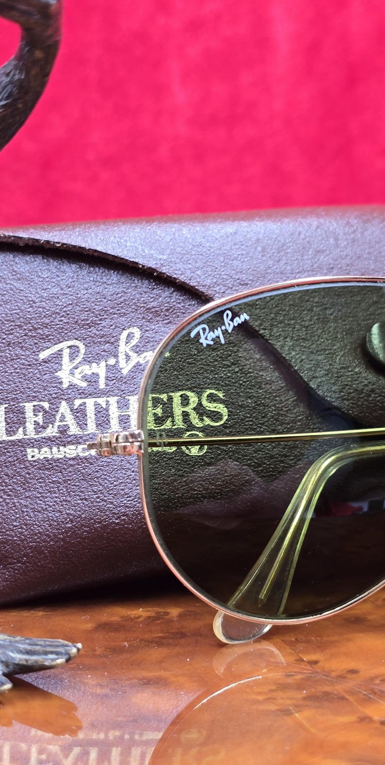 May include: Close-up of Ray-Ban sunglasses with a gold frame and dark lenses. The Ray-Ban logo is visible on the lens. A brown leather case with the Ray-Ban logo is in the background. The sunglasses are a classic aviator style.